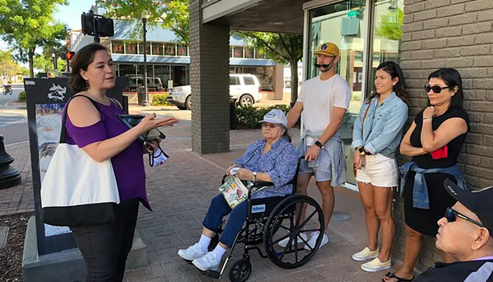 A group of people including an individual in a wheelchair attentively listens to a woman who appears to be giving a tour or presentation outdoors