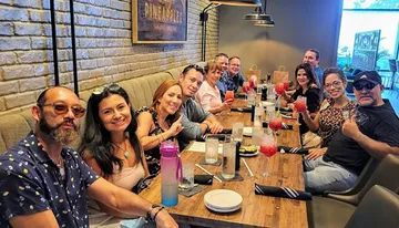 A group of people is smiling and posing for a photo while sitting at a long dining table with drinks, suggesting a social gathering or a celebration.