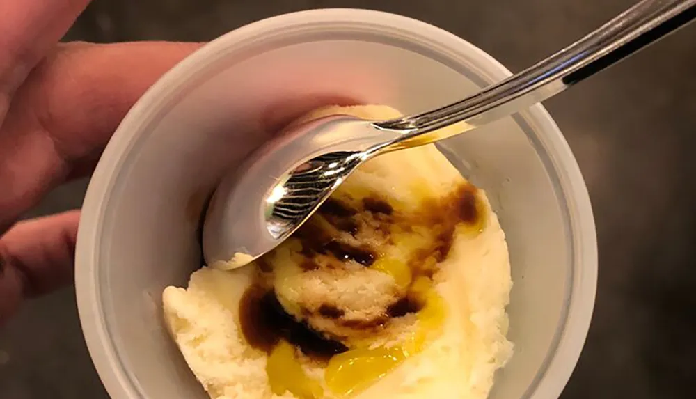 The image shows a close-up view of a person holding a spoon and a plastic cup containing a dessert that resembles mashed potatoes with gravy captured in a way that might be intentionally misleading
