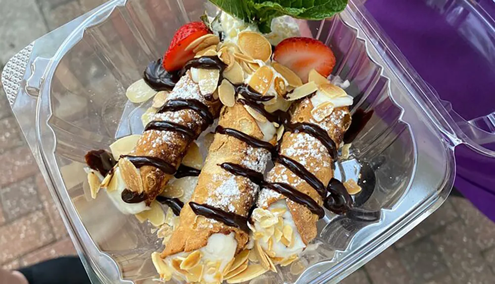 The image features a pair of cannolis topped with powdered sugar drizzled with chocolate sauce garnished with sliced strawberries and almond flakes and served in a clear plastic container