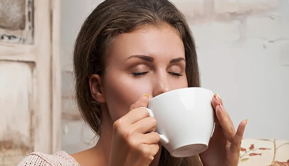 A woman is savoring the aroma of a beverage from a large white cup with her eyes closed in appreciation