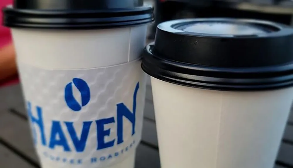 Two takeaway coffee cups with black lids are placed on a table with the closest one displaying a logo that includes the text HAVEN COFFEE ROASTERS