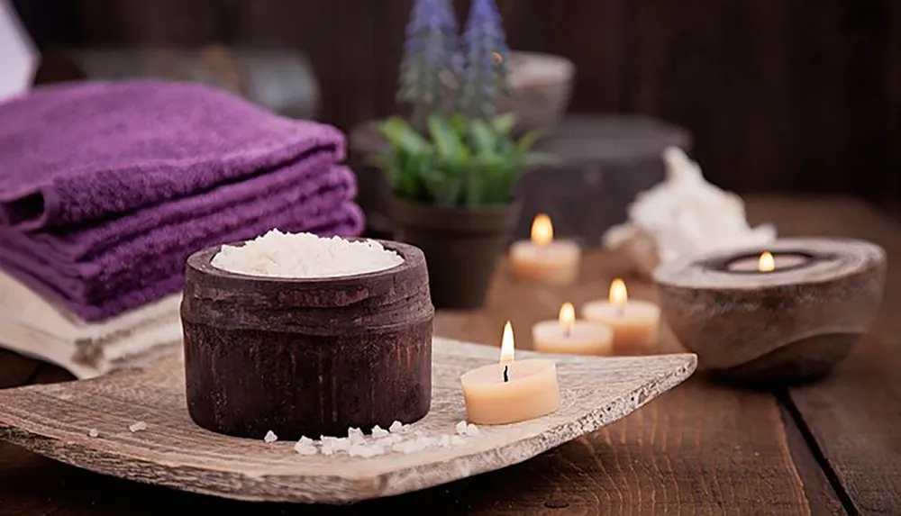 The image depicts a serene spa setting with lit candles a bowl of bath salts a stack of purple towels and a plant creating an atmosphere of tranquility and relaxation