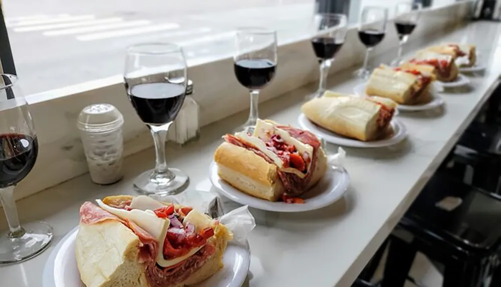 A row of sandwiches and glasses of red wine are neatly arranged on a bar counter facing a window