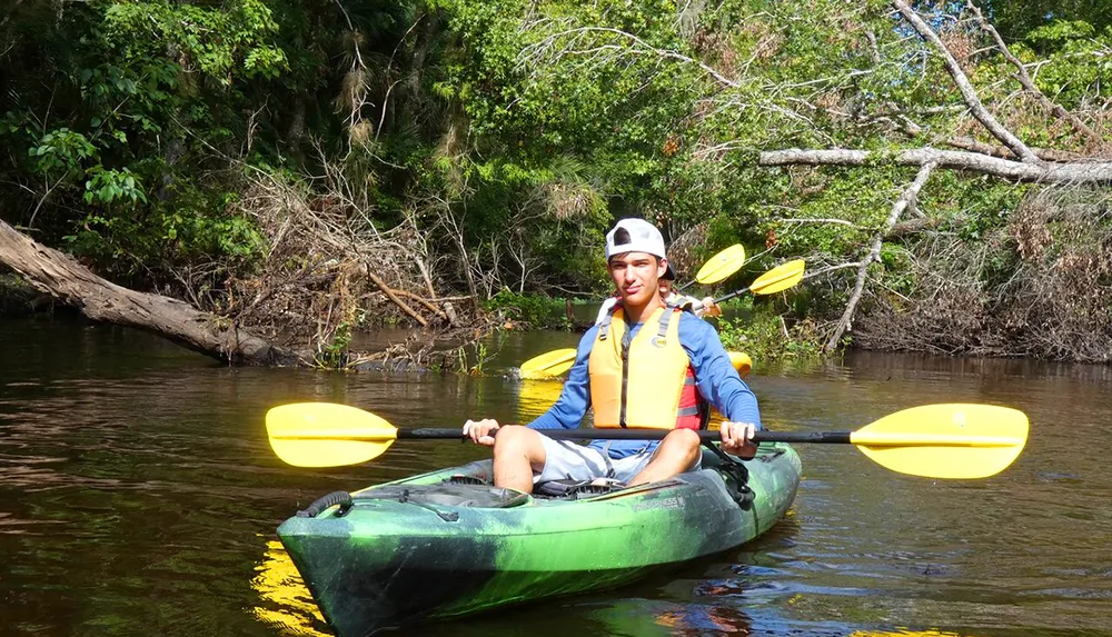 A person wearing a life jacket and cap is kayaking on a calm river surrounded by lush greenery under a clear sky