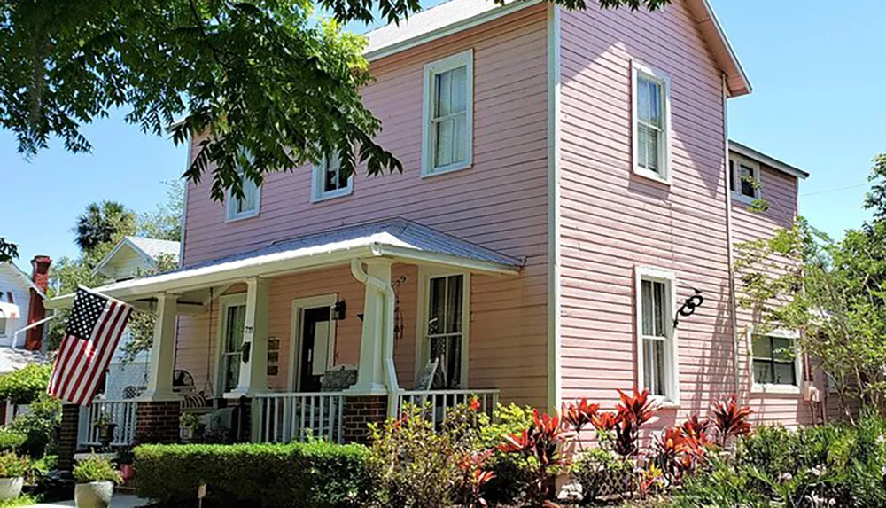 The image shows a two-story pink house with white trim featuring a front porch with an American flag surrounded by a green lawn and various plants on a sunny day