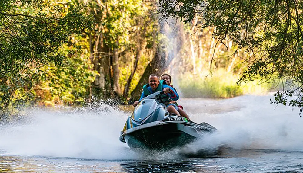 Two people are joyfully riding a jet ski at high speed creating a splash of water surrounded by a natural landscape