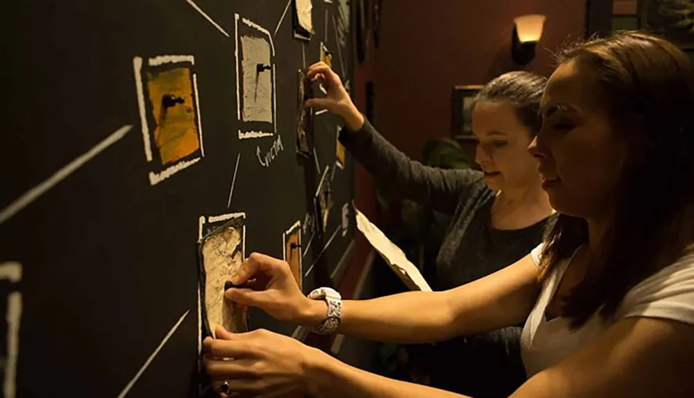 Two people are engaged in an interactive wall activity arranging pieces on a chalkboard surface illuminated by warm lighting
