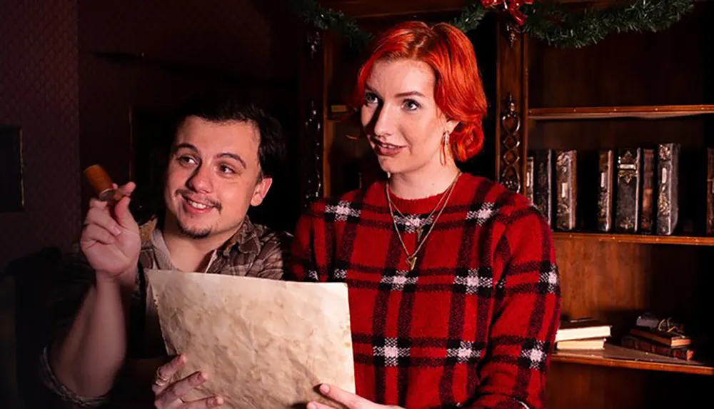 A man holding a cigar and a piece of paper smiles as he looks off-camera while a woman with red hair looks at the viewer both dressed in cozy attire against a background suggestive of a festive old-fashioned interior