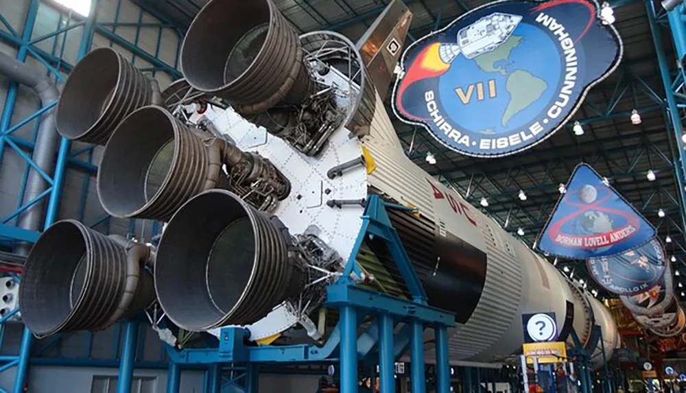 The image features the powerful engine section of a Saturn V rocket displayed in a museum with mission insignia banners hanging above