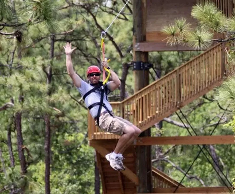 A person wearing a red helmet and safety harness is zip-lining through a forested area smiling and waving at the camera