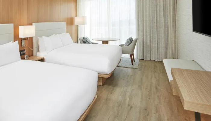 The image shows a modern and neatly organized hotel room with two large beds a TV table with chairs and a wooden-panel backdrop