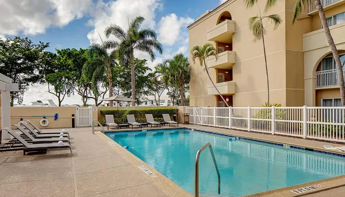 The image shows an outdoor swimming pool area with sun loungers at an apartment complex enclosed by a white fence with palm trees and a building in the background