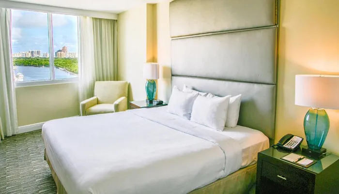 The image displays a bright hotel room with a large bed modern furnishings and a view of a waterway through a window