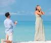 A man is kneeling on a sandy beach holding an engagement ring towards a surprised woman with her hands over her face