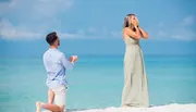 A man is kneeling on a sandy beach holding an engagement ring towards a surprised woman with her hands over her face.