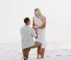 A man is kneeling on a sandy beach holding an engagement ring towards a surprised woman with her hands over her face