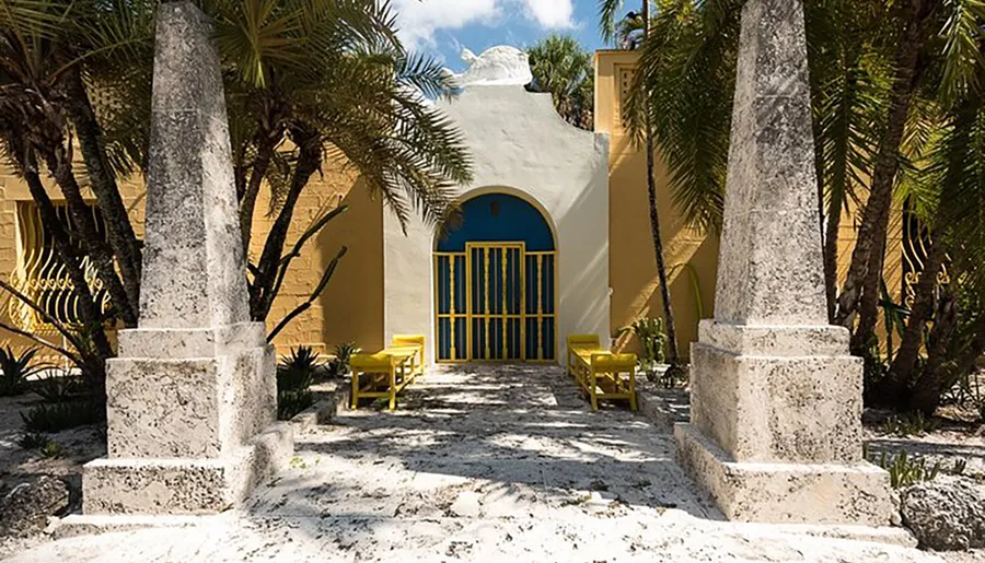 A symmetrical view of a bright yellow building with an arched blue doorway flanked by white stone pillars and tropical foliage, accented by two yellow chairs on a sunny day.