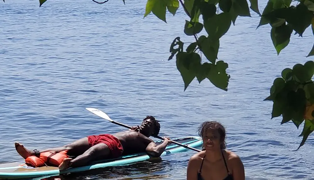 A person is lying on a paddleboard relaxing with a paddle in hand while another individual is standing in water nearby framed by green leaves against a backdrop of calm water