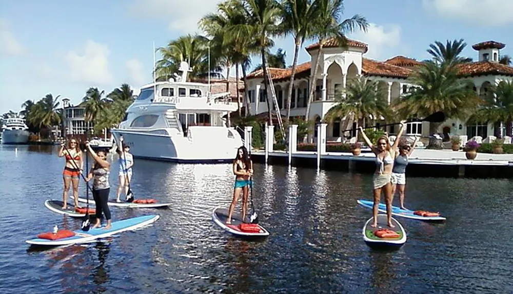 A group of people is enjoying stand-up paddleboarding in a sunny calm waterway with a luxurious mansion and a yacht in the background