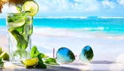 A refreshing glass of mojito alongside a pair of sunglasses is laid out on a table with a tropical beach backdrop.
