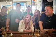 A group of smiling people are enjoying drinks together at a bar, with some wearing green beads, suggesting a festive or celebratory occasion.
