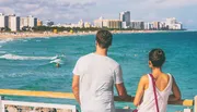 A couple is enjoying the view of a vibrant beach scene with surfers and a city skyline in the background.