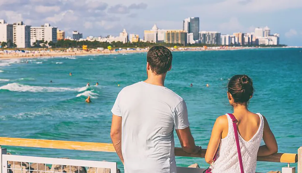 A couple is enjoying the view of a vibrant beach scene with surfers and a city skyline in the background