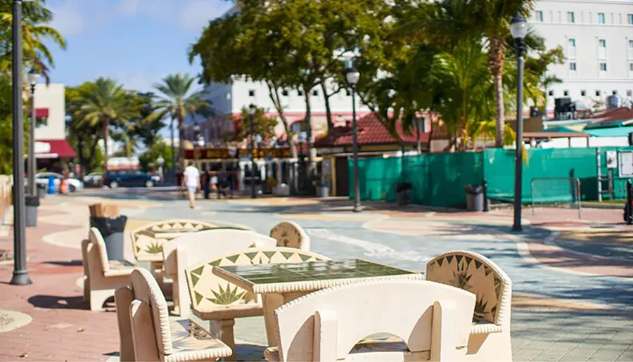 Sunlight bathes a pedestrian street lined with palm trees and equipped with sculptural benches, creating a relaxed urban scene.
