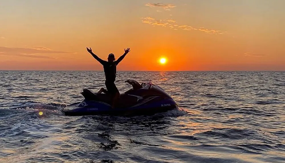 A person strikes a joyful pose atop a jet ski on a wavy sea against a stunning sunset