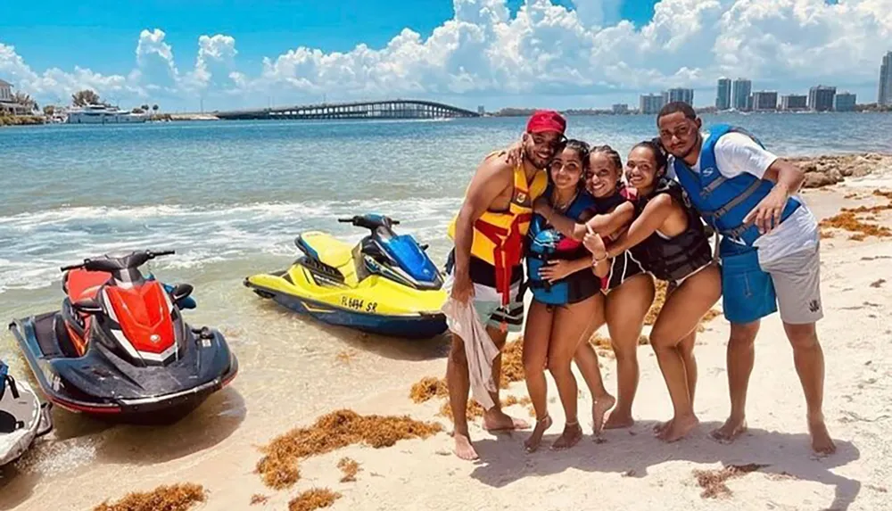 A group of five people wearing swimwear and life jackets are smiling for a photo on a sunny beach with two jet skis nearby and a bridge and skyline in the background