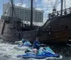 Two individuals are riding jet skis on the water near a docked replica of an old sailing ship