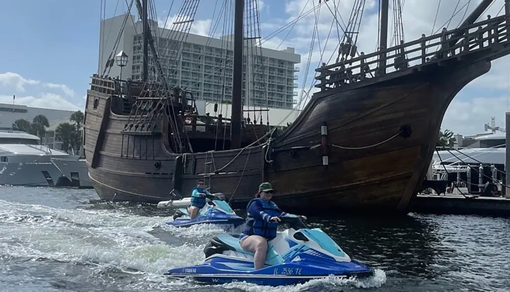 Two individuals are riding jet skis on the water near a docked replica of an old sailing ship