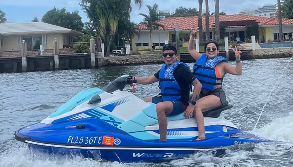 Two people are happily jet skiing on the water waving at the camera with a tropical coastline in the background