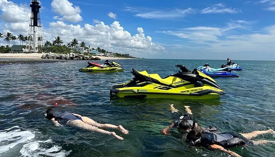 A group of people are snorkeling near a shoreline dotted with palm trees, with multiple jet skis floating nearby, under a partly cloudy sky.