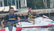 Two people wearing life vests are smiling and riding separate jet skis near a waterfront property.