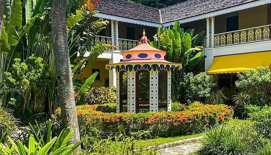 The image shows a vibrant garden with a whimsical gazebo in front of a brightly painted building surrounded by tropical vegetation.