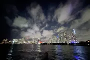 The image shows a nighttime city skyline viewed across a body of water, with a dramatic cloudy sky overhead.