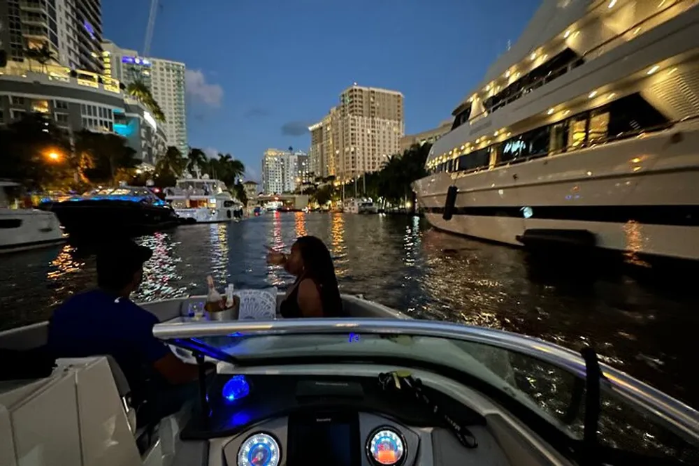Two people are enjoying an evening boat ride amidst other boats and illuminated city buildings in the background