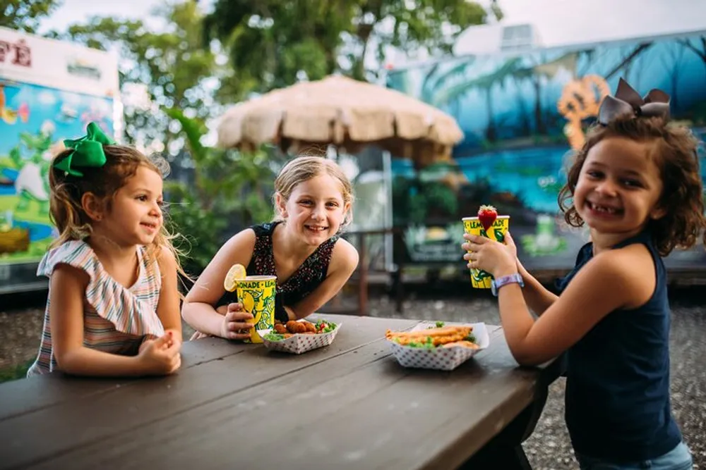 Three young girls are smiling and sitting at an outdoor table with snacks and beverages enjoying each others company