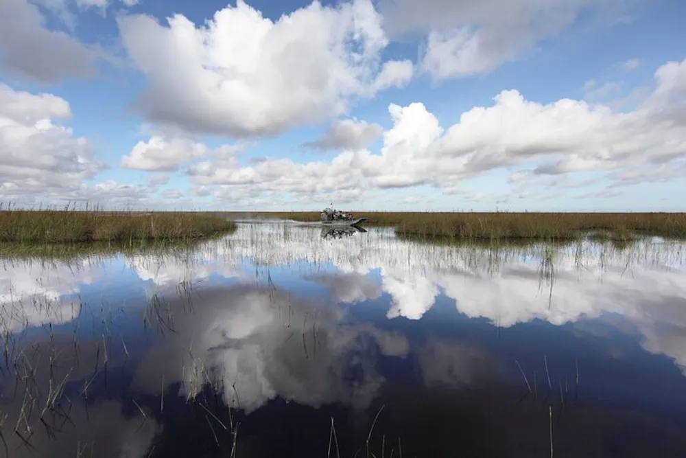 An airboat glides across a calm water surface reflecting clouds in the Everglades