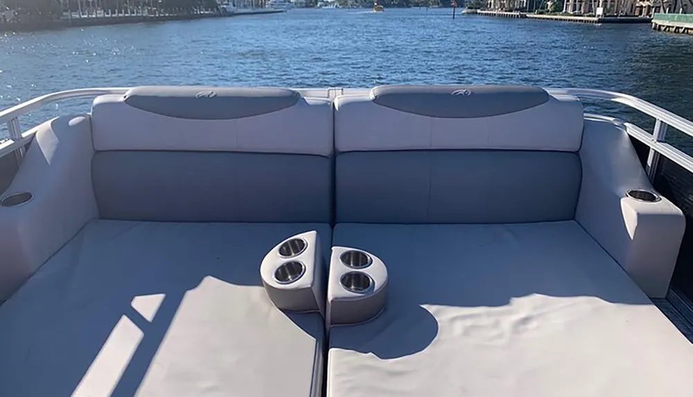 This is a photograph of a boats stern with cushioned seating and cup holders overlooking calm water with a shoreline in the distance