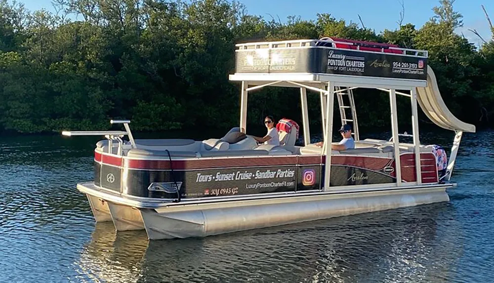 A pontoon boat with passengers is cruising near a lush shoreline under bright sunlight