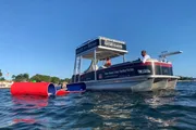 A pontoon boat is floating near an overturned red kayak with a person in the water reaching out to the vessel under a clear blue sky.