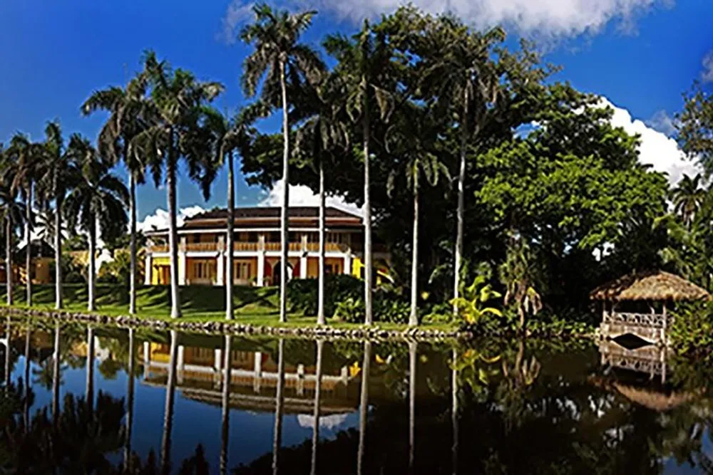 The image depicts a tranquil scene of a colonial-style mansion set among tall palm trees reflected in the still waters of a pond with a small thatched hut on the right