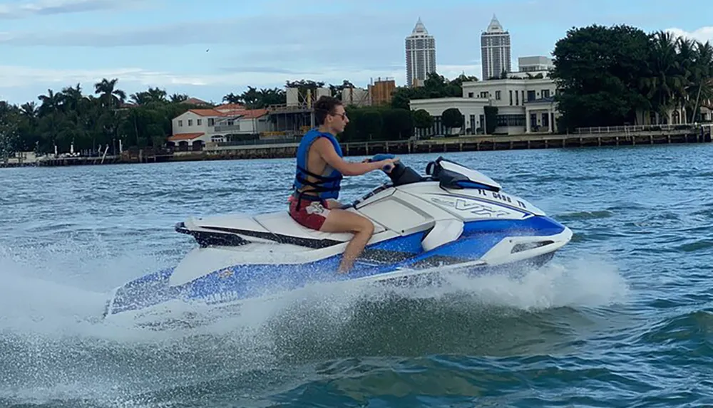 A person is riding a jet ski on the water with coastal buildings in the background