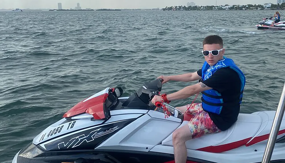 A person is seated on a stationary jet ski on the water wearing a life vest and sunglasses with other watercraft and the shoreline visible in the background