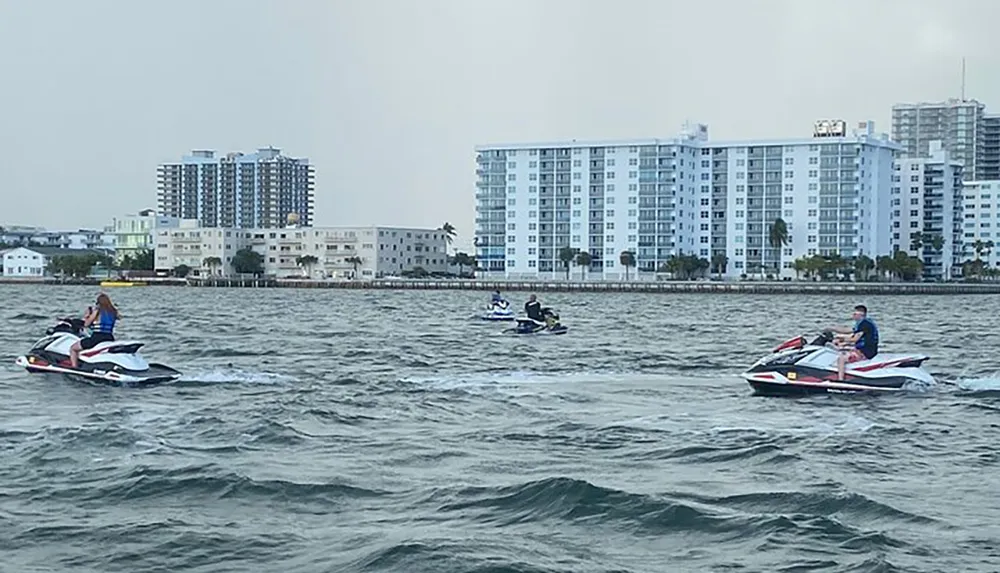 Individuals are riding jet skis on a choppy water surface with coastal city buildings in the background