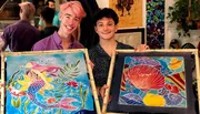 Two individuals are holding up colorful paintings of fish, with smiles on their faces, possibly having completed an art class or workshop.