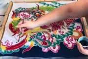 An artist's hand is shown painting a vibrant and detailed phoenix on fabric stretched over a frame.
